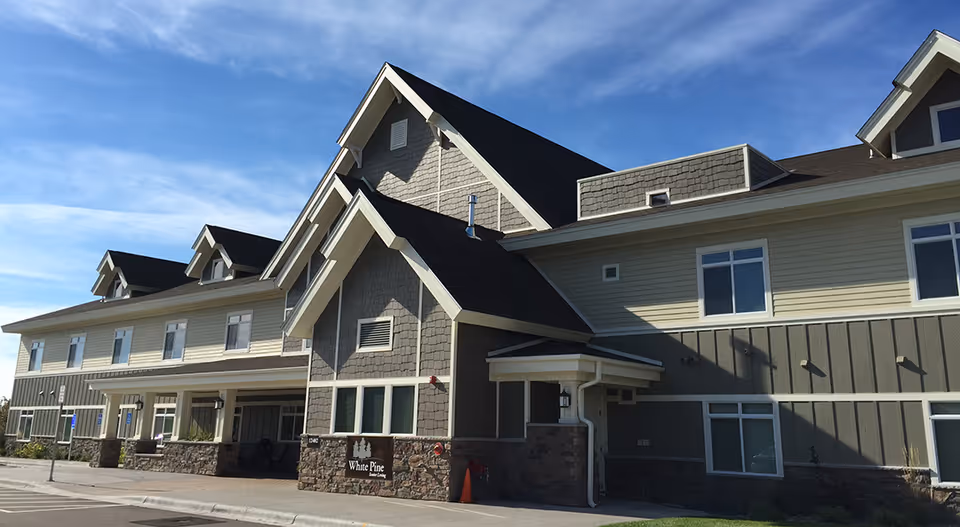 Front exterior of a two-story senior living facility with pitched roofs and a stone entry displaying a 'White Pine' sign.
