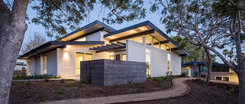 Exterior view of a modern single-story building at dusk with large windows and a sloped roof, surrounded by trees and a curved dirt pathway leading to the entrance.
