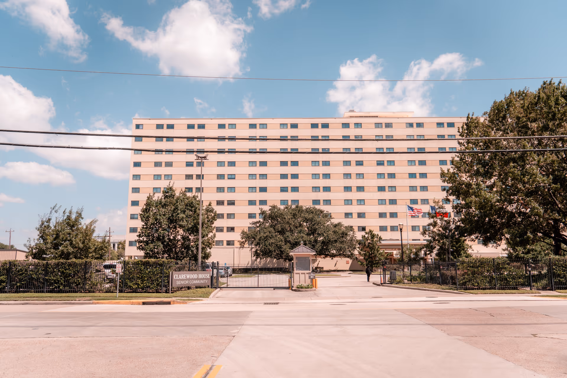 Exterior front view of a large multi-story senior living building with a gated entrance, trees, and flags.