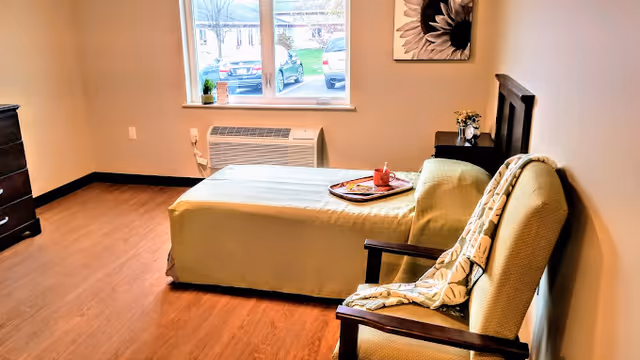 A cozy bedroom with a single bed covered in a light green bedspread, a wooden chair with a patterned blanket draped over it, a small nightstand with a flower vase, a window showing parked cars outside, and a framed flower artwork on the wall.