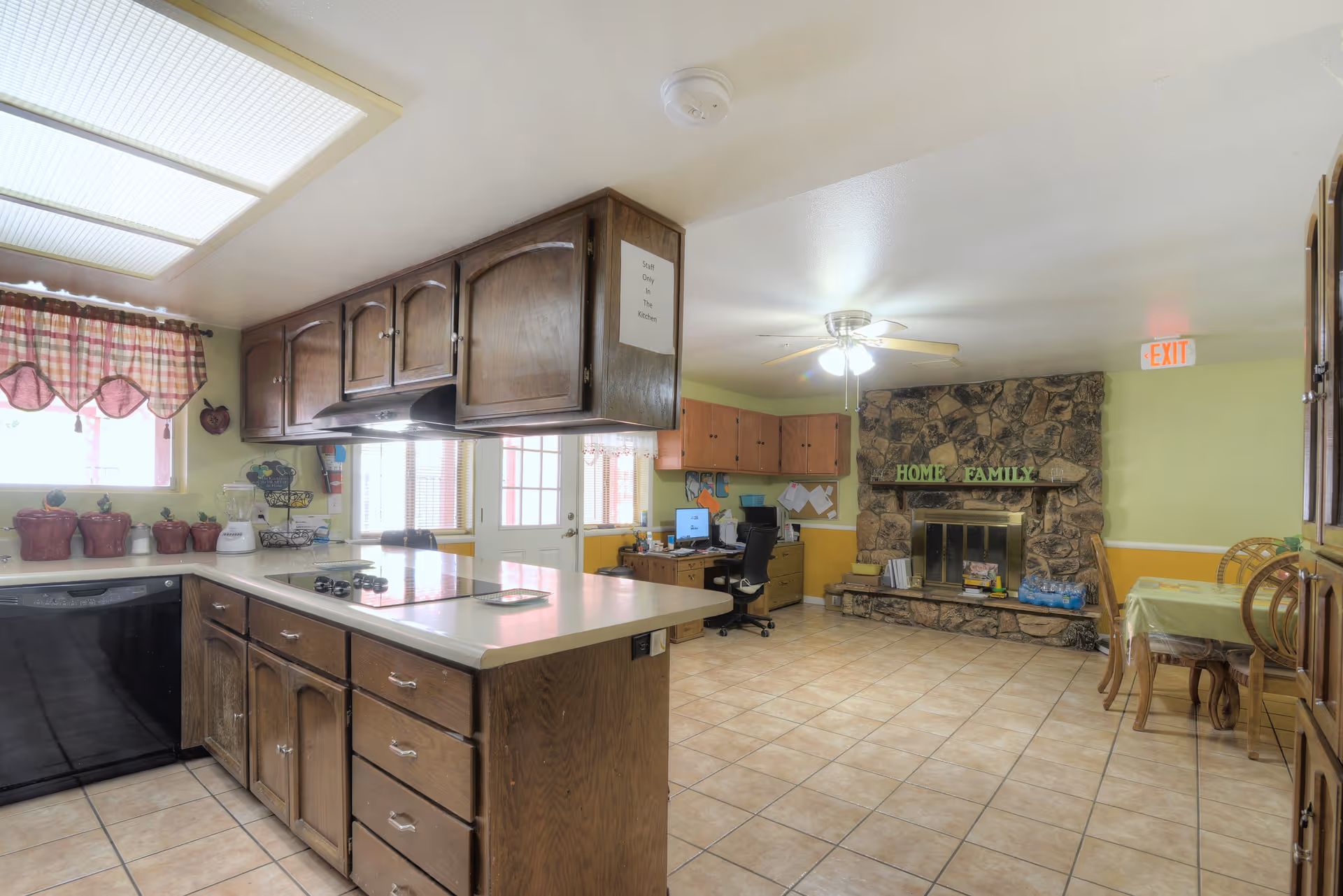 Interior view of a combined kitchen and living area in an assisted living facility. The kitchen features wooden cabinets, a countertop with a stovetop, and various kitchen items including red canisters and a blender. The living area has a stone fireplace with decorative letters spelling 'HOME FAMILY' on the mantel, a ceiling fan with lights, a desk with a computer, and a dining table with chairs. The walls are painted light green and yellow, and there is an exit sign above a door in the background.