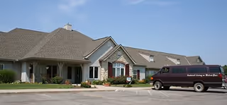 Exterior view of a single-story senior living facility building with a pitched roof, stone and siding facade, and a maroon van parked in front with the text 'Assisted Living at Manor Place' on its side.