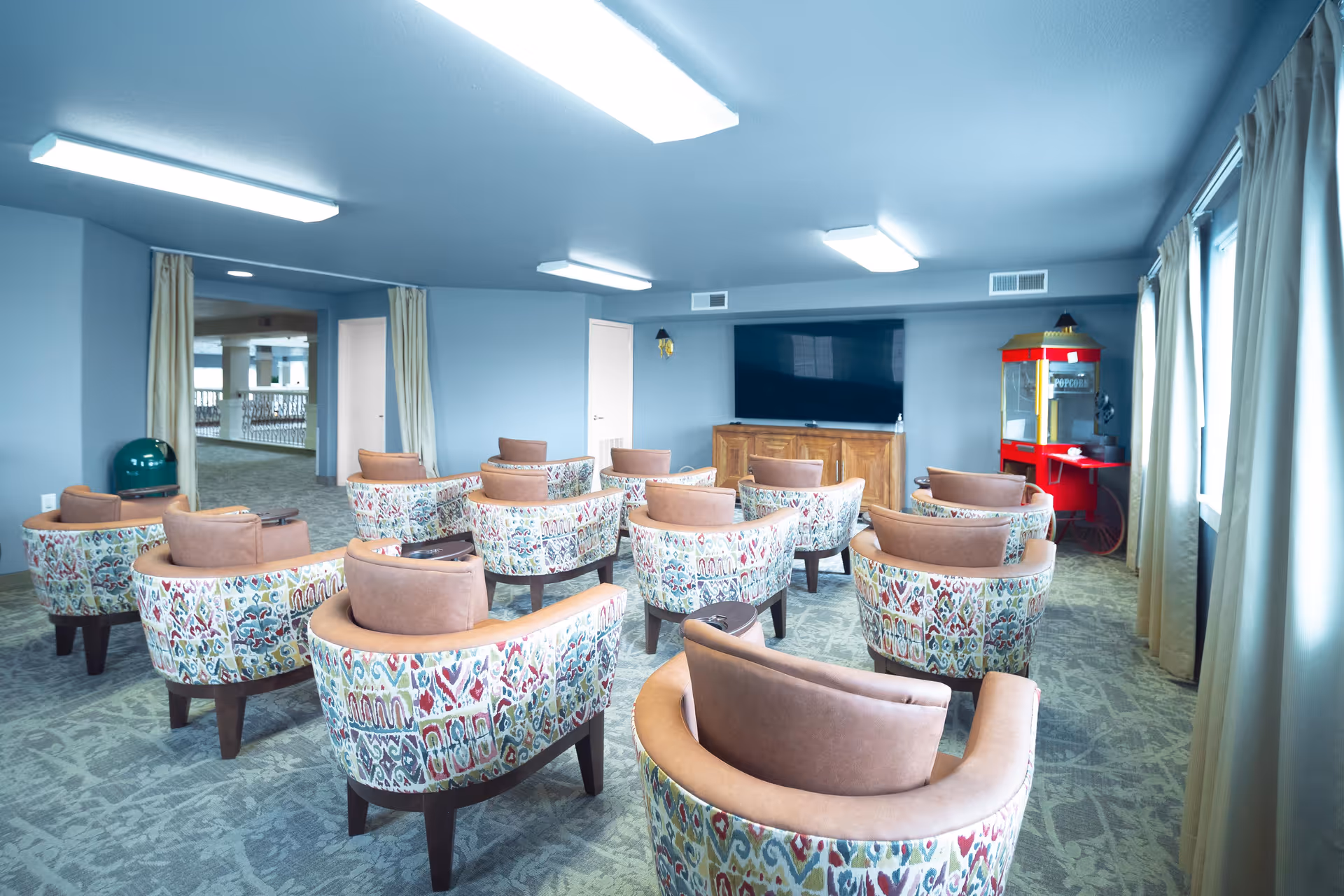 A cozy common area with patterned armchairs arranged in rows facing a large flat-screen TV mounted on a wooden cabinet. To the right, there is a red popcorn machine. The room has light blue walls, carpeted floor, and large windows with beige curtains allowing natural light to enter.
