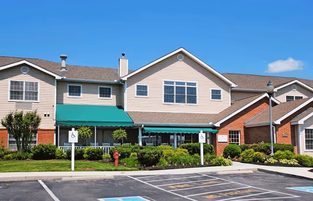 Exterior view of a two-story senior living facility building with beige siding and red brick accents. The building has green awnings over the windows and a well-maintained garden with shrubs and small trees in front. There are two handicap parking signs visible in the parking lot under a clear blue sky.