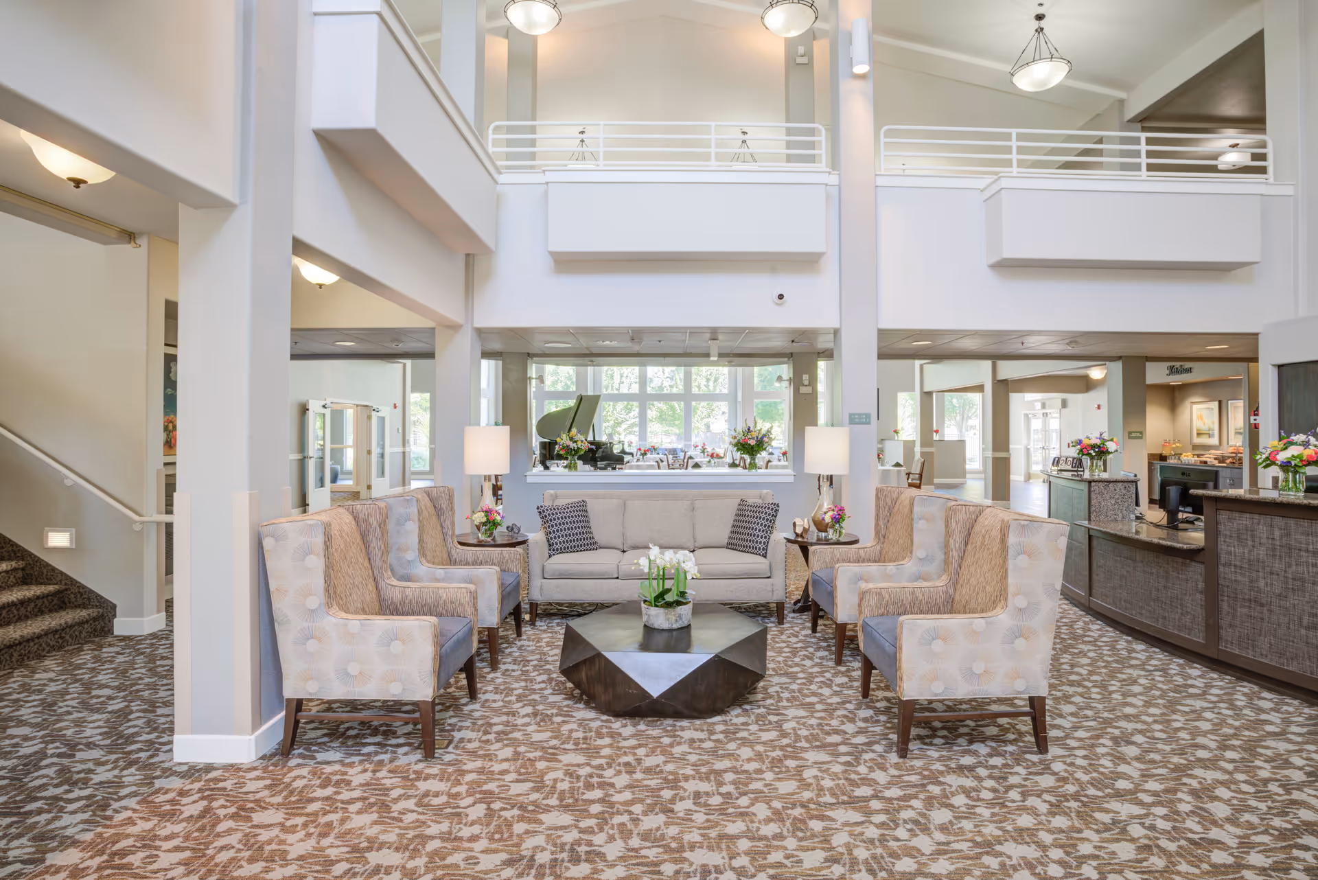 A spacious and well-lit senior living facility common area with a seating arrangement consisting of a central sofa, six armchairs, and a modern geometric coffee table. The room features high ceilings, large windows letting in natural light, patterned carpet, and decorative lamps on side tables. In the background, there is a reception desk and a grand piano near the windows.