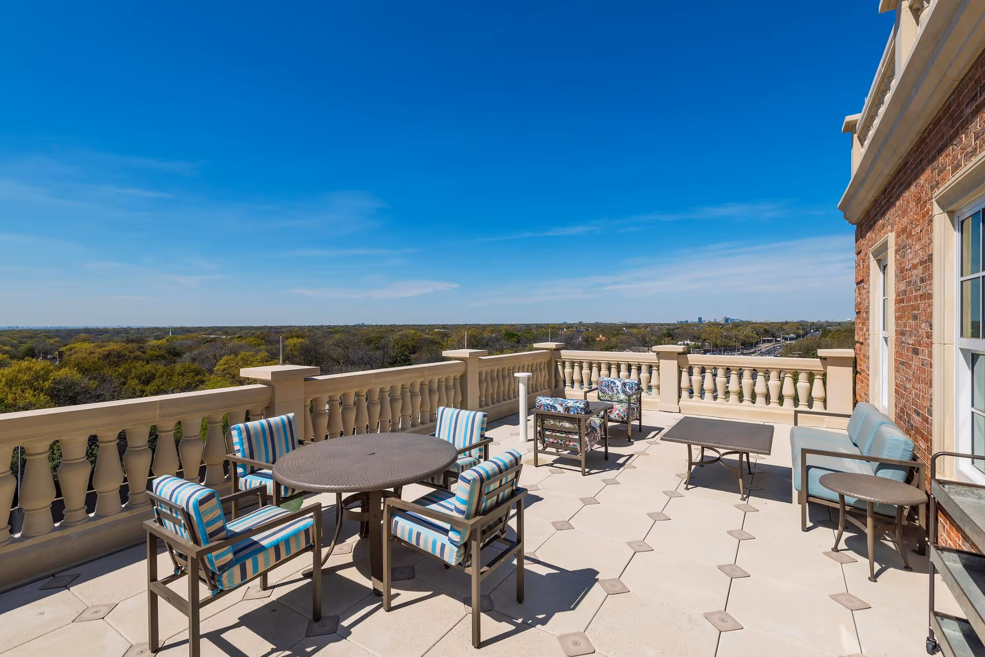 Rooftop terrace with tables, striped cushioned chairs, and a balustrade overlooking treetops and a distant skyline.