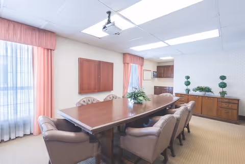 A well-lit conference room with a long wooden table surrounded by eight cushioned chairs. The room has large windows with sheer white curtains and pink drapes, a ceiling-mounted projector, and a wooden cabinet with decorative plants on top.