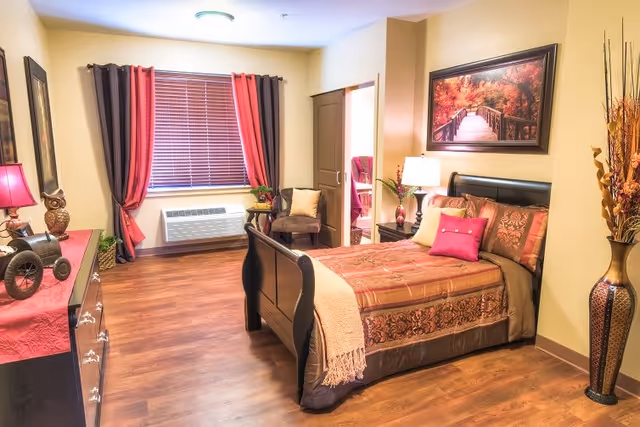 A warmly decorated bedroom with a wooden bed featuring patterned bedding and multiple pillows. There is a nightstand with a lamp and decorative items beside the bed. A framed picture of a wooden bridge in autumn hangs on the wall above the bed. The room has wooden flooring, a window with red and brown curtains, a chair in the corner, and a dresser with decorative items including an owl figurine and a lamp. A tall vase with dried branches stands in the corner near the bed.