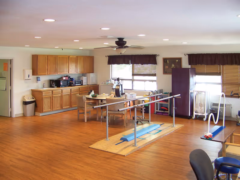 A spacious room with wooden flooring featuring a kitchenette area with wooden cabinets, a microwave, and a coffee maker. In the center, there is a table with four chairs and various items on it. The room also contains physical therapy equipment including parallel bars and balance beams. Large windows with brown valances allow natural light to enter the room.