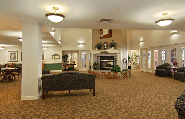 Spacious senior living facility common area with patterned carpet, multiple seating arrangements including sofas and chairs, a central fireplace with plants and decorative items, wall sconces, ceiling lights, and large windows with French doors allowing natural light.