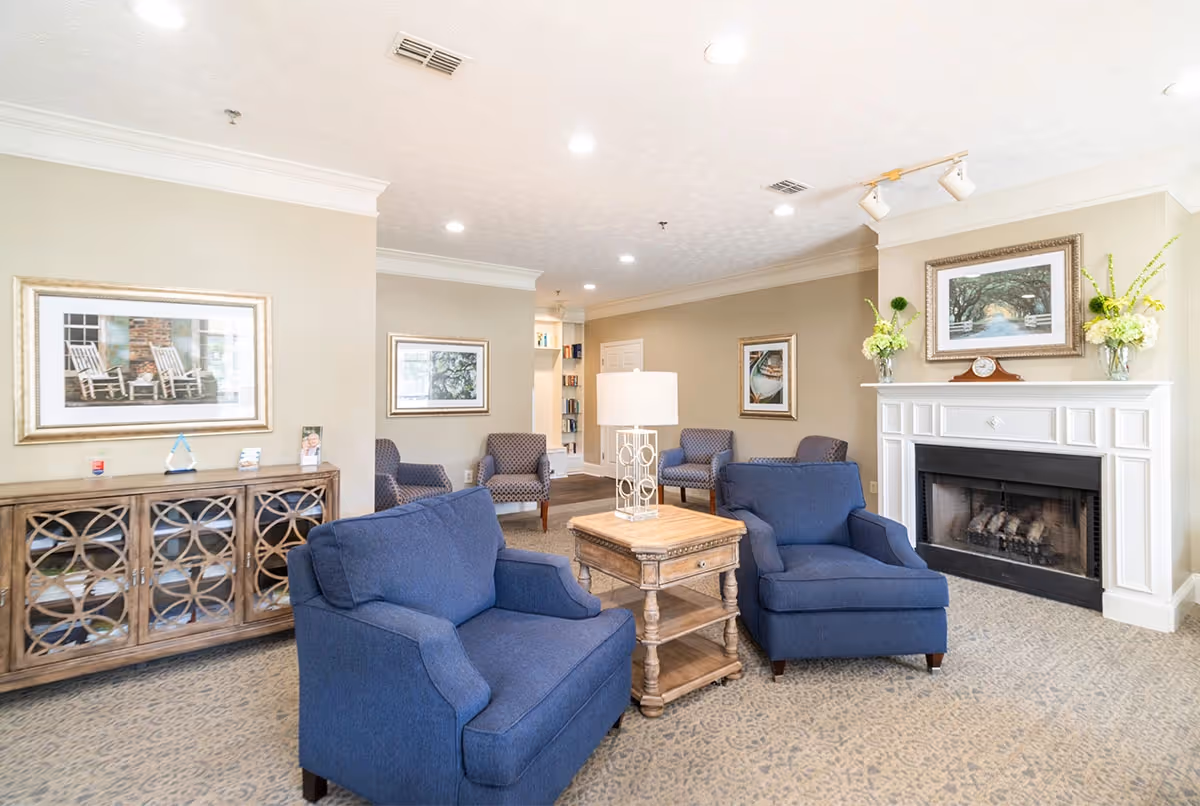 A cozy senior living community lounge featuring two blue armchairs around a wooden side table with a lamp, a white fireplace with a clock and floral arrangements on the mantel, framed artwork on beige walls, and additional seating with patterned chairs in the background.