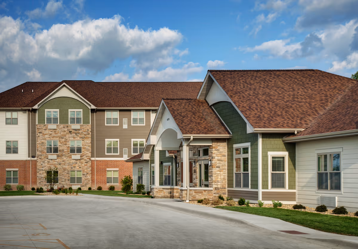 Exterior view of a senior living facility building with a combination of stone, brick, and siding on the facade. The building has multiple windows and a covered entrance with a peaked roof. The sky is partly cloudy with blue patches visible.