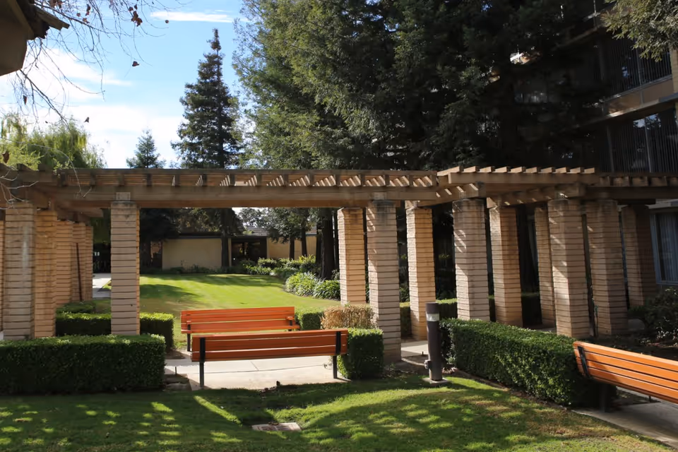 A landscaped outdoor courtyard with a wooden pergola supported by brick columns, benches, hedges, and a grassy lawn.