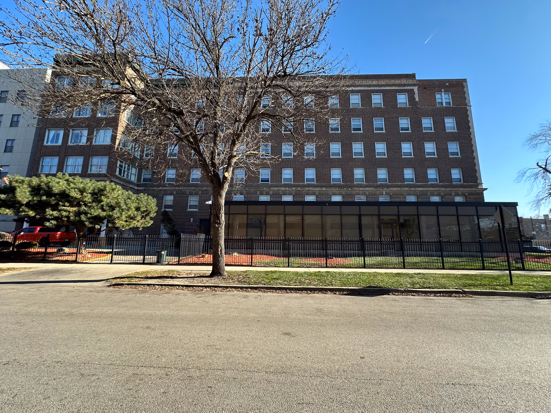Exterior view of a multi-story brick building with many windows, a large leafless tree in front, a fenced grassy area, and a clear blue sky.