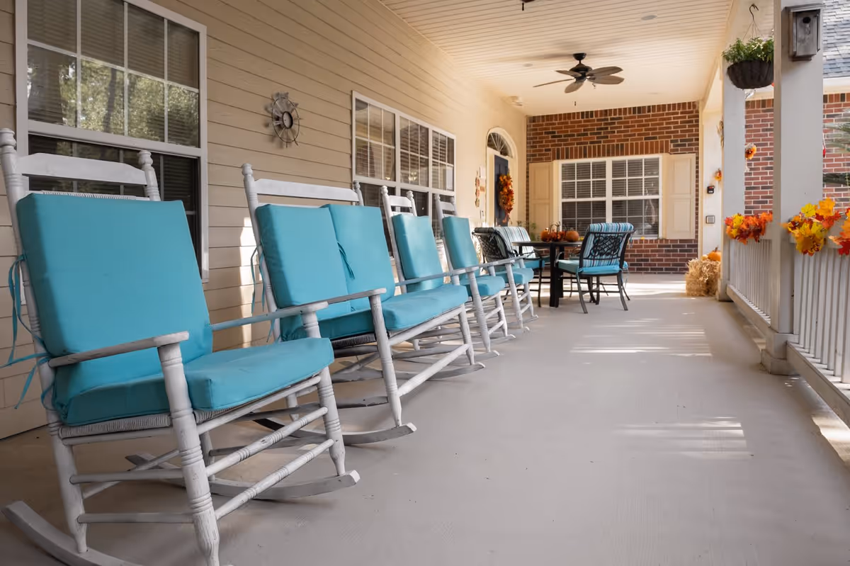 A covered porch area with a row of white rocking chairs featuring turquoise cushions on the left side. The porch has beige siding, windows with white frames, a ceiling fan, and a table with chairs in the background. Autumn-themed decorations with orange and yellow leaves adorn the railing and walls.