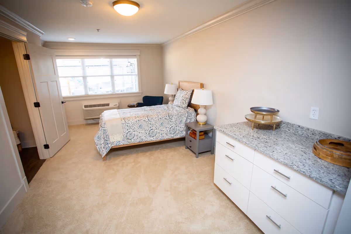 Bright furnished bedroom with a single bed, nightstand and armchair by a window and a dresser with a granite top.