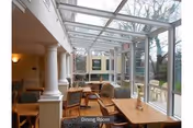 A bright dining room with large windows and a glass ceiling allowing natural light to fill the space. The room features wooden tables and chairs arranged neatly, with some cushioned seating. White columns and a view of outdoor greenery are visible through the windows.