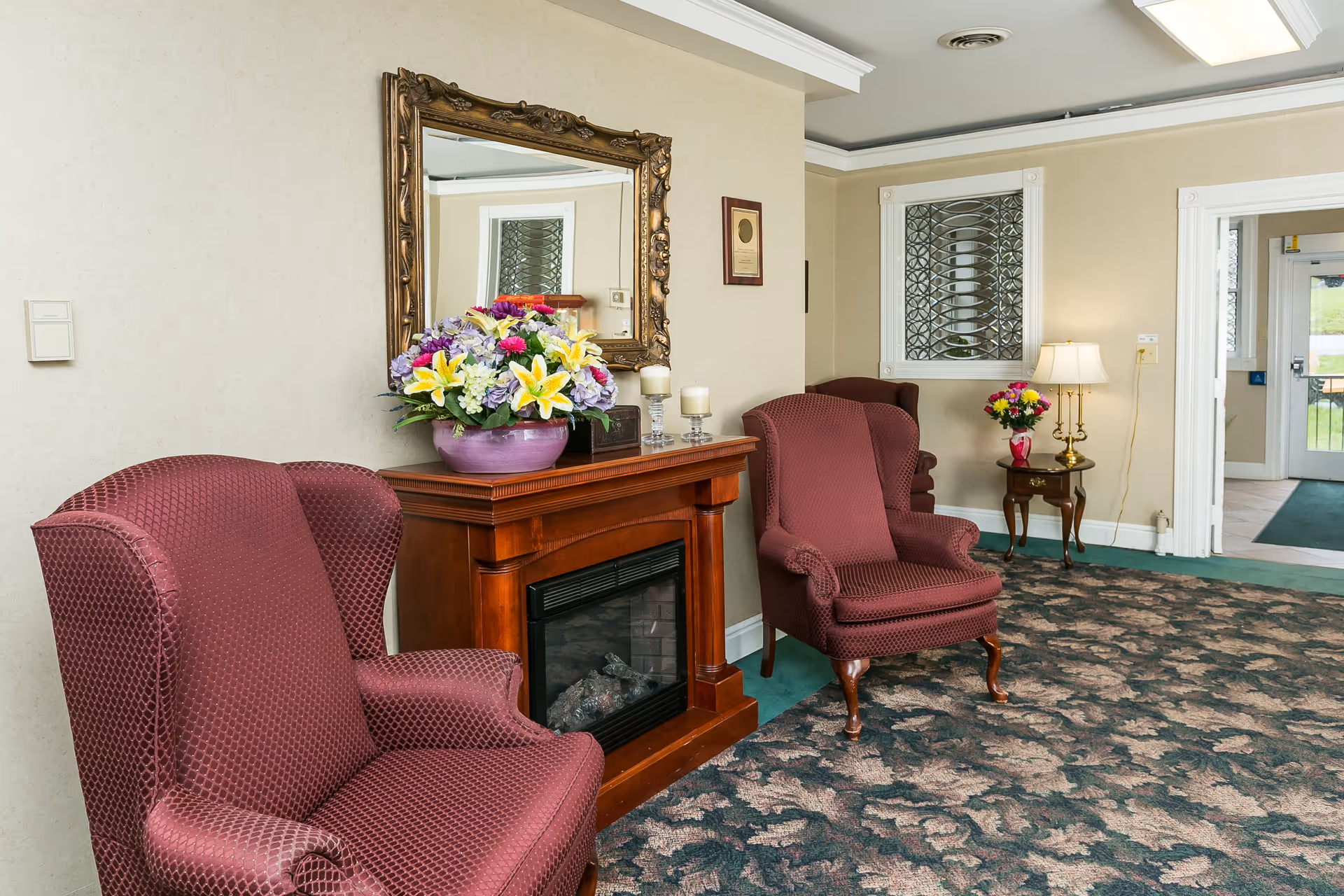 Lobby seating area with two upholstered armchairs, a wooden mantel with a floral arrangement and mirror, and a side table with a lamp.