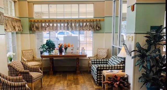 A cozy sitting area with a wooden table holding flower vases in front of large windows with valance curtains. Surrounding the table are cushioned chairs with plaid and floral patterns. There are plants and a lamp adding to the warm, inviting atmosphere.
