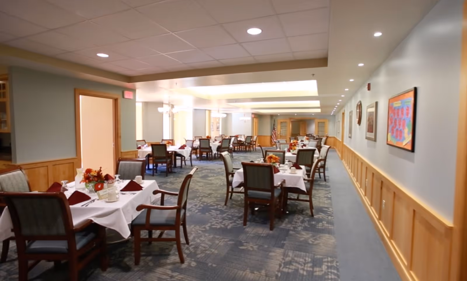 A spacious dining room in a senior living community with multiple tables covered in white tablecloths, each set with cups, napkins, and floral centerpieces. The room has soft lighting, carpeted floors with a floral pattern, wooden paneling on the walls, and framed artwork hanging on the right wall.