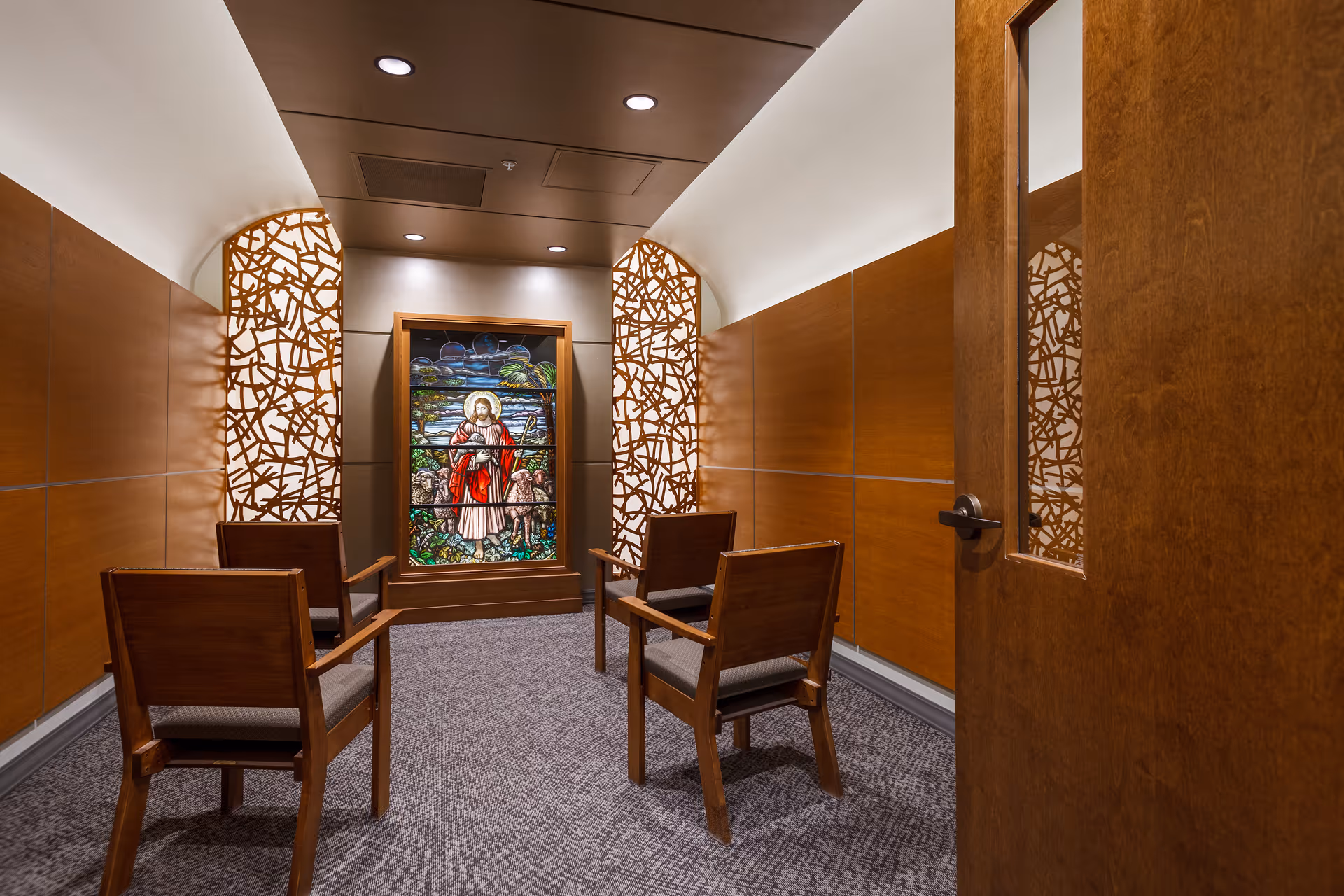 A small, quiet room with four wooden chairs arranged in a square facing a stained glass window depicting a religious figure holding a staff, surrounded by sheep and nature. The walls are paneled with wood, and decorative wooden lattice panels flank the stained glass. The room has a carpeted floor and recessed ceiling lights.