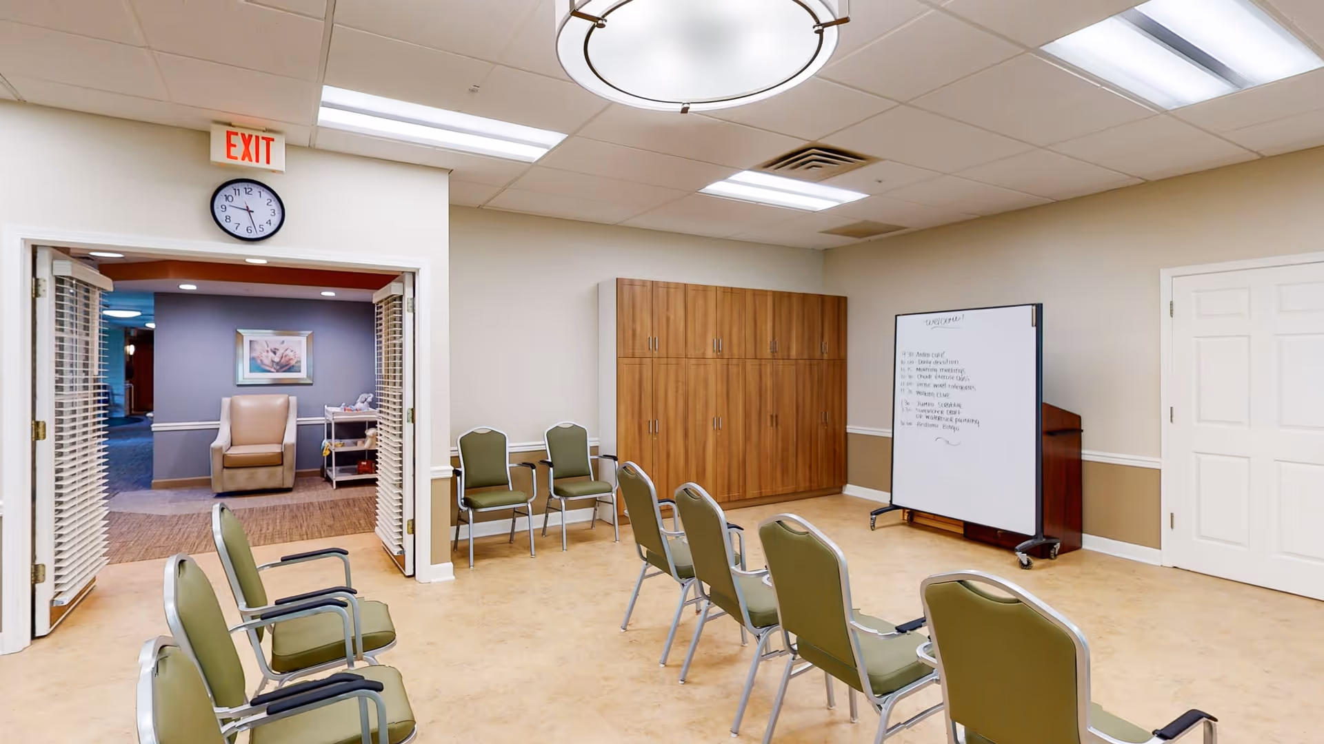 A meeting or activity room with green chairs arranged in rows facing a whiteboard on wheels. There is a wooden cabinet against the wall and a clock above the doorway showing 10:10. The room has beige walls with a white chair rail and a tan floor. Through the open double doors, a lounge area with an armchair and framed artwork is visible.