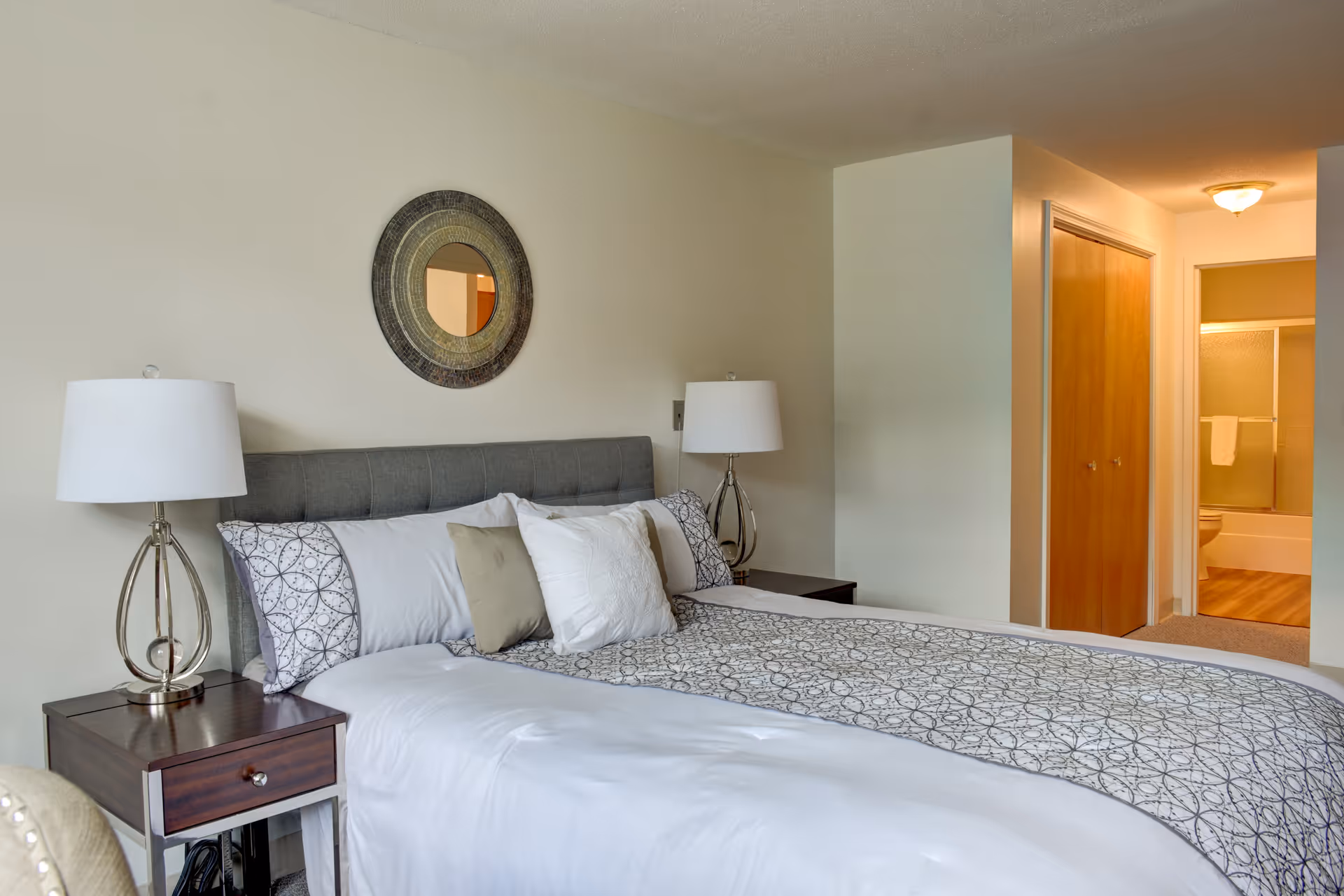 Well-appointed bedroom with a made bed, two bedside tables and lamps, a round mirror above the headboard, and an open doorway leading to a bathroom.
