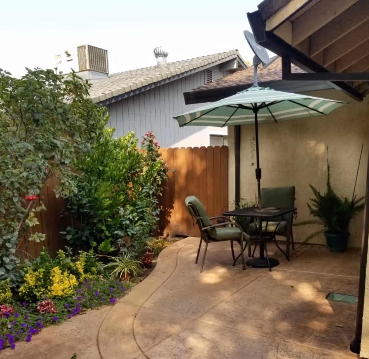 Small enclosed patio with a table, chairs and a striped umbrella next to plants and a wooden fence.