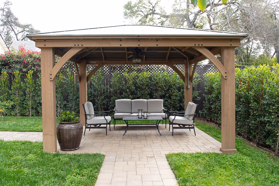Wooden gazebo in a garden with patio furniture including a sofa, chairs, and a coffee table.