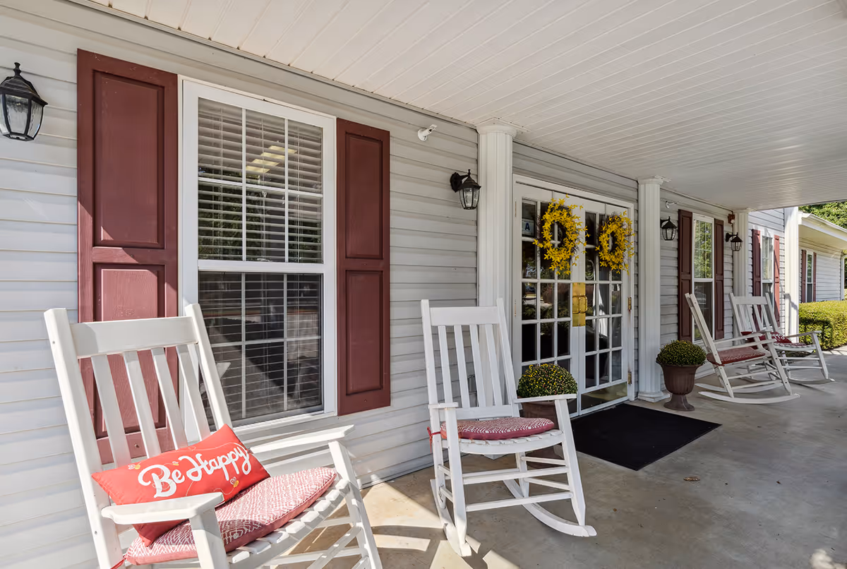 Covered porch area with white rocking chairs featuring red cushions and a red pillow with the words 'Be Happy'. The porch has white siding with maroon shutters on the windows, two white columns, black wall-mounted lanterns, and double glass doors decorated with yellow wreaths. There are also potted plants near the entrance.