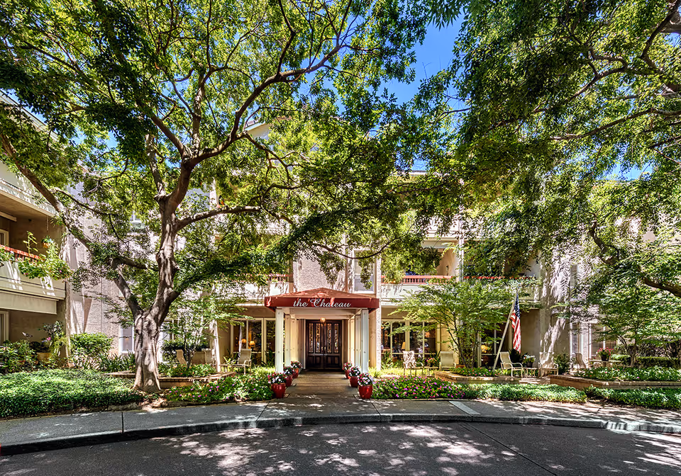 Front exterior view of a senior living facility named 'the Chateau' with a covered entrance, surrounded by lush green trees and landscaped flower beds. There are chairs and an American flag near the entrance.