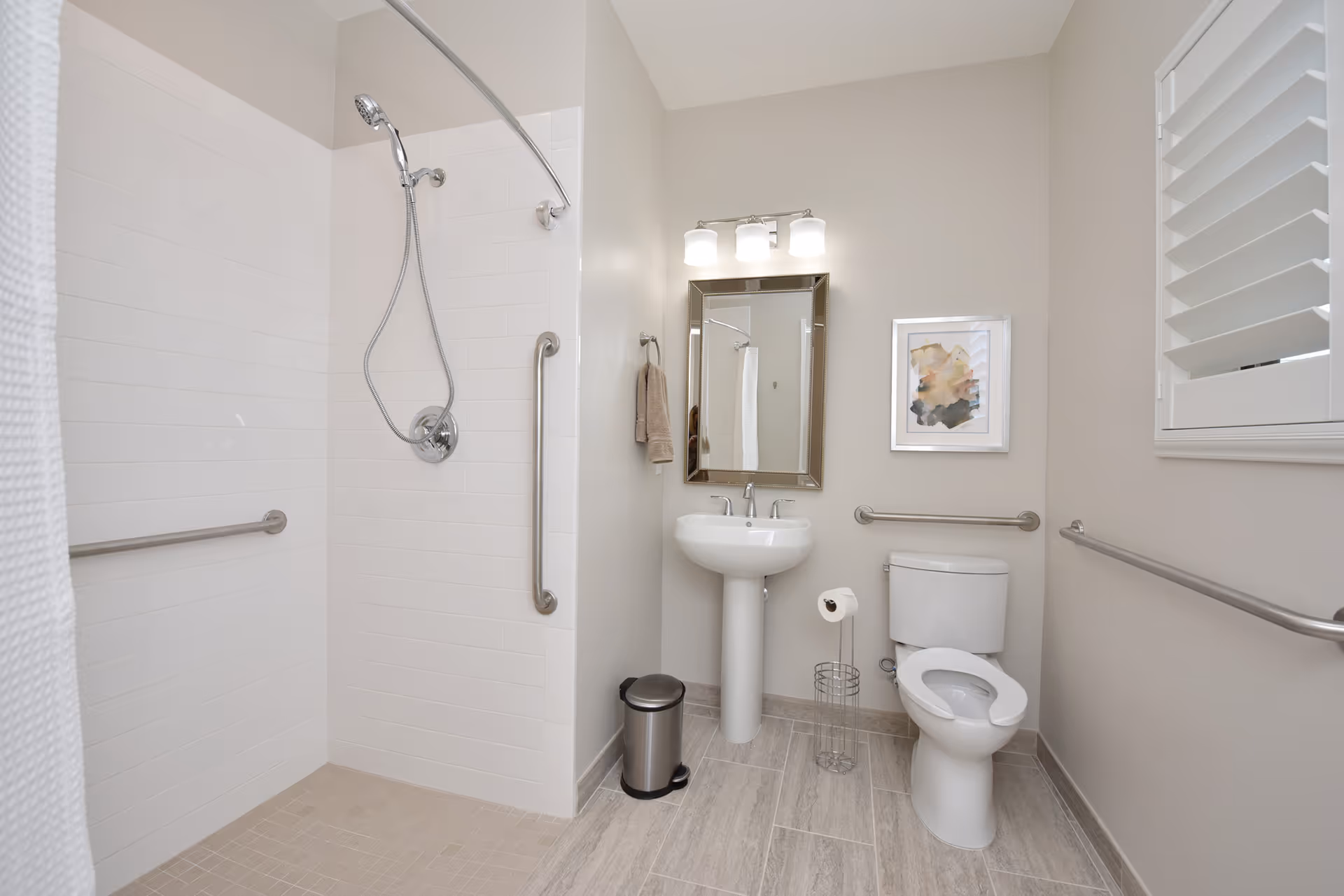 A clean, modern bathroom with a walk-in shower featuring a handheld showerhead and grab bars. The bathroom also includes a pedestal sink with a mirror and light fixture above it, a toilet with a raised seat, a toilet paper holder, a small trash can, and a framed abstract artwork on the wall. The walls are painted light beige, and the floor is tiled in a light wood-like pattern.