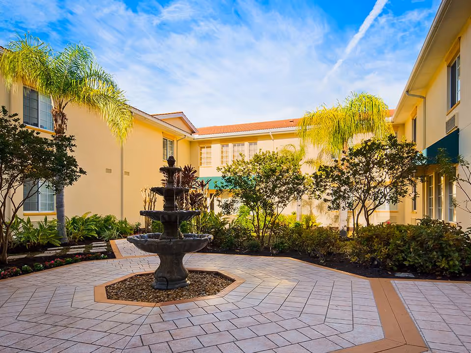 Sunny courtyard with a central tiered fountain, palm trees and landscaped gardens surrounded by a beige two-story building.