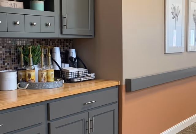 A section of a kitchen or kitchenette area with gray cabinets and drawers, a wooden countertop, a tray with syrup bottles, a plant, and a wire basket holding napkins and cups. The wall has a mosaic tile backsplash and two framed floral artworks are visible on the adjacent wall.