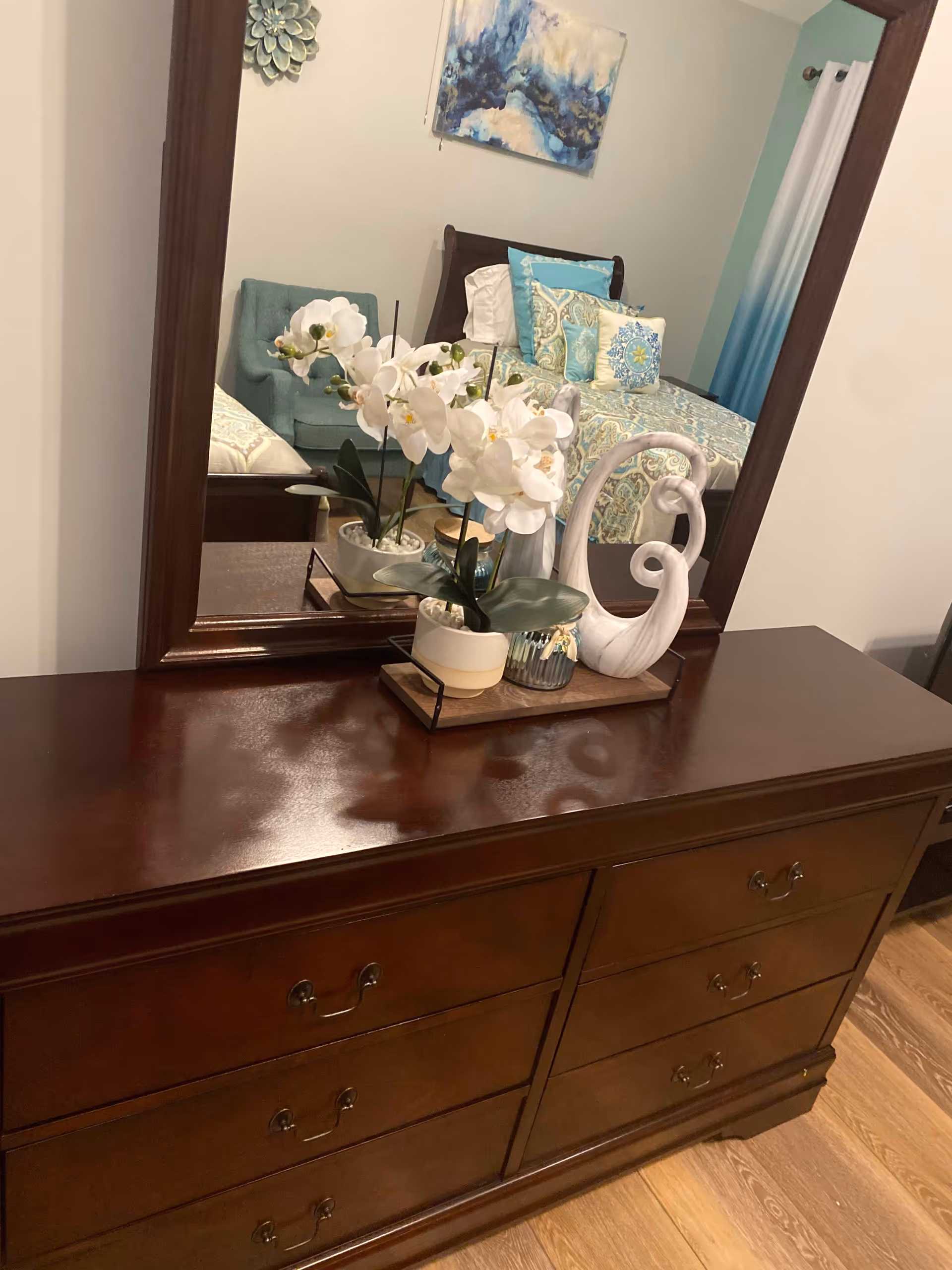 A wooden dresser with six drawers and a large mirror above it. On top of the dresser, there is a decorative tray holding a white orchid plant, a small candle holder, and a white ornamental sculpture. The mirror reflects a bed with patterned bedding, several pillows, a teal armchair, and a piece of abstract wall art.