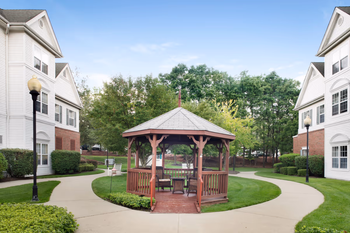 A wooden gazebo sits in a landscaped courtyard with curved walkways between two white multi-story residential buildings.