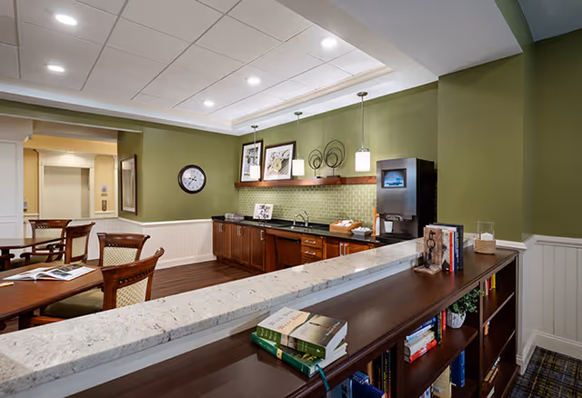 Interior view of a common area in a senior living facility with a green accent wall, a countertop with books and decorative items, wooden cabinets, a coffee machine, framed artwork, a wall clock, and a dining table with chairs.