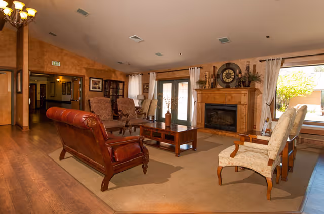 A cozy living room area with wooden flooring and beige walls. The room features a brown leather sofa, two upholstered armchairs, a wooden coffee table, and a fireplace with decorative items on the mantel. Large windows with white curtains allow natural light to brighten the space.