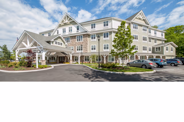 Exterior view of a multi-story senior living facility building with a covered entrance, stone and siding facade, several windows, landscaped greenery, and a parking lot with cars under a partly cloudy sky.