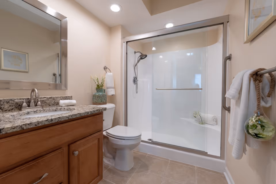 A clean and modern bathroom featuring a granite countertop with a sink, a large mirror above the sink, a toilet, and a glass-enclosed shower with a handheld showerhead. The bathroom has beige walls, tiled floor, and decorative elements including a vase with flowers and framed artwork on the walls. Towels are neatly hung on towel racks.