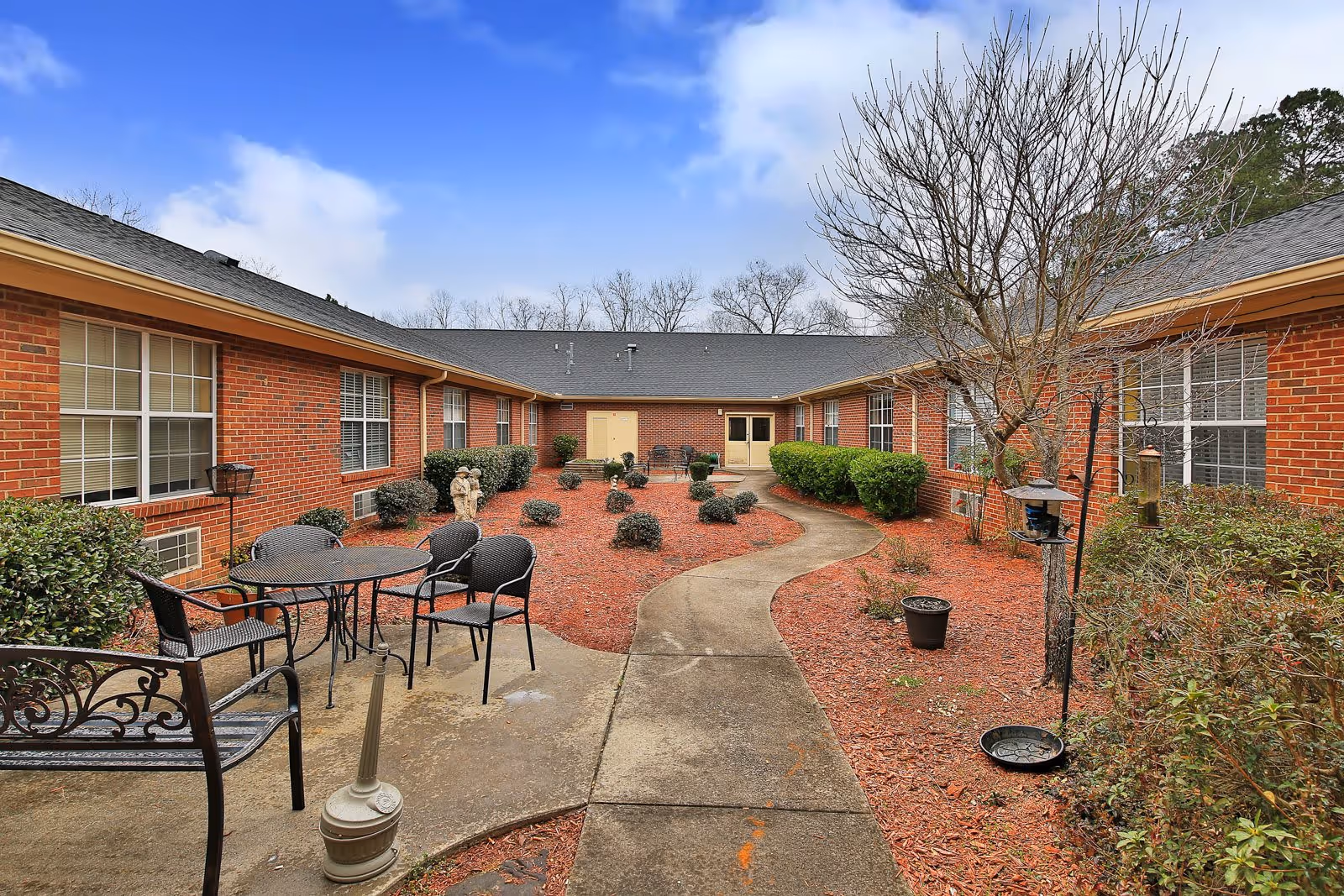 Outdoor courtyard area at Manchester Court Assisted Living and Memory Care with a curved concrete walkway, surrounded by red mulch and various shrubs and small trees. There are black metal chairs and a table on the left side, along with a bench and a bird feeder on the right. The courtyard is enclosed by a single-story brick building with multiple windows and a cloudy blue sky overhead.