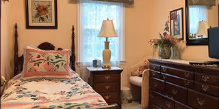 A cozy bedroom with a single bed featuring floral bedding and a decorative pillow. Next to the bed is a wooden nightstand with a lamp. A window with sheer curtains allows natural light into the room. Across from the bed is a wooden dresser with a mirror above it, a vase with flowers, and a teddy bear on a chair beside the dresser. The walls are decorated with framed artwork.
