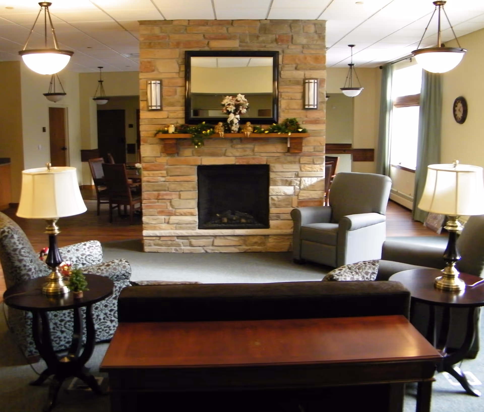 Common living room with a central stone fireplace, armchairs, side tables with lamps, and a mirror above the mantel.