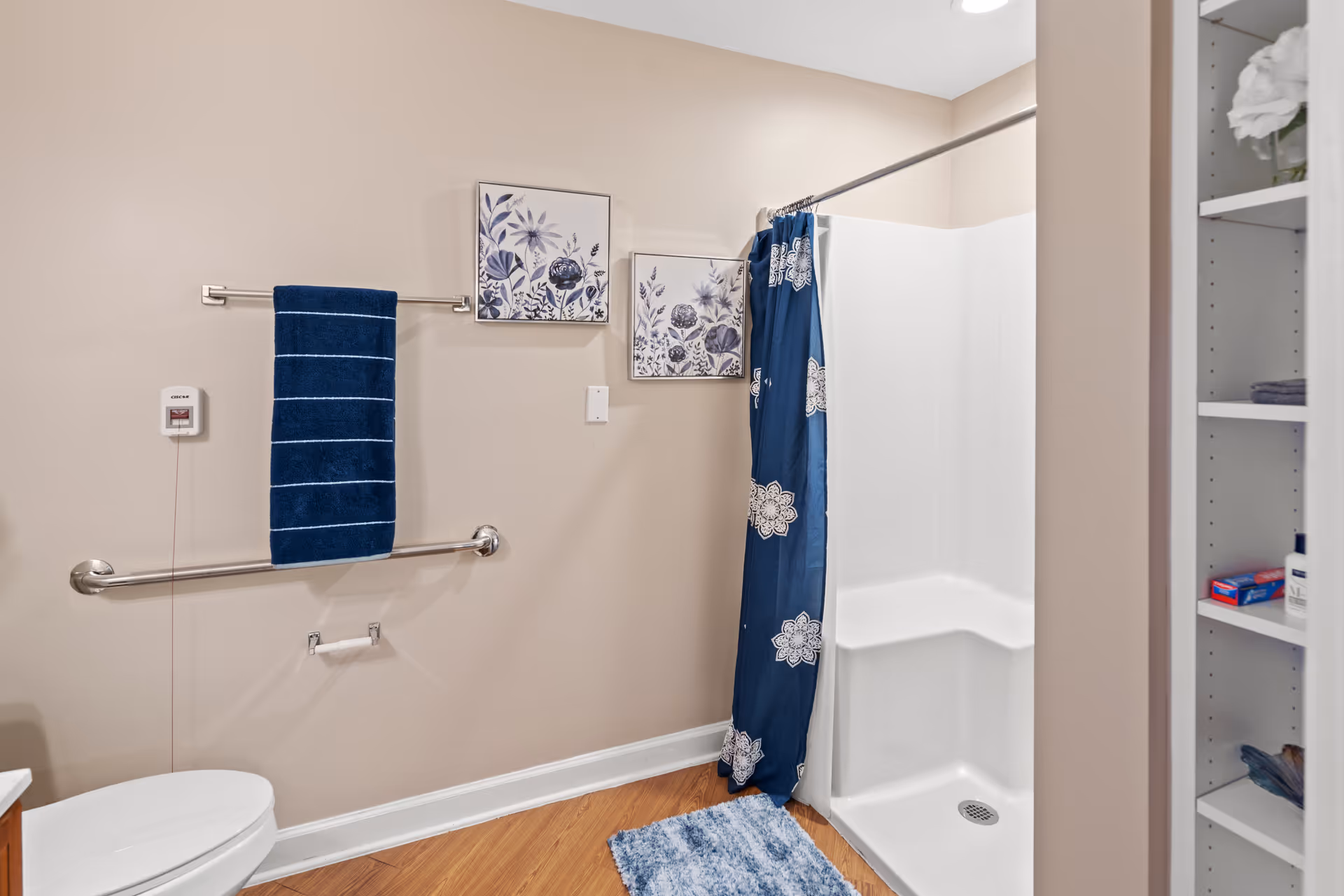 A clean and accessible bathroom featuring a white shower with a built-in seat and a blue floral shower curtain. The bathroom has beige walls with two framed floral artworks, a blue towel hanging on a metal bar, a toilet, and a shelving unit with toiletries and towels. The floor is wooden with a blue bath mat in front of the shower.