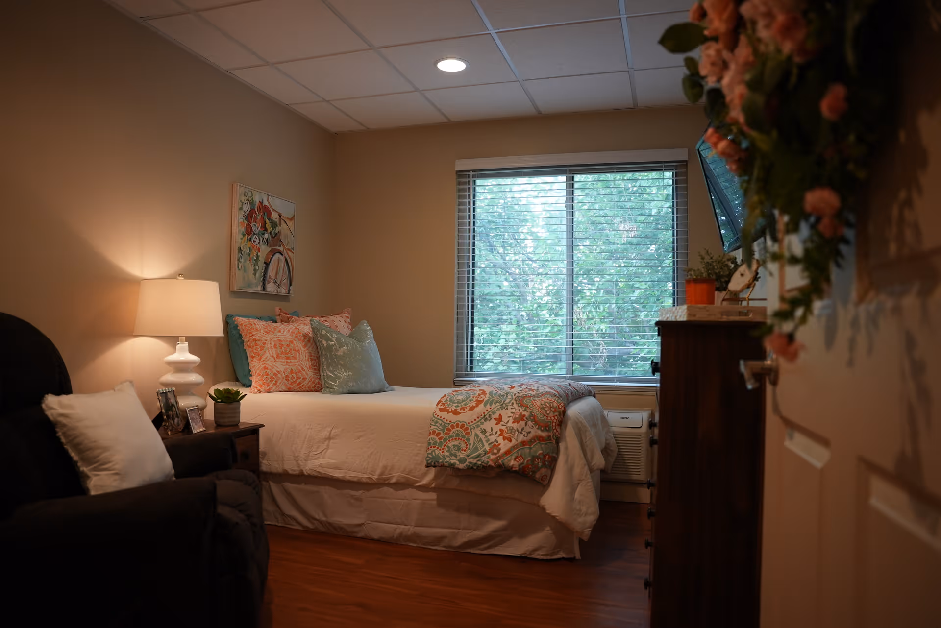 A cozy bedroom with a made bed topped with patterned pillows and a throw, a lamp on a nightstand, armchair, window with blinds, and a dresser.