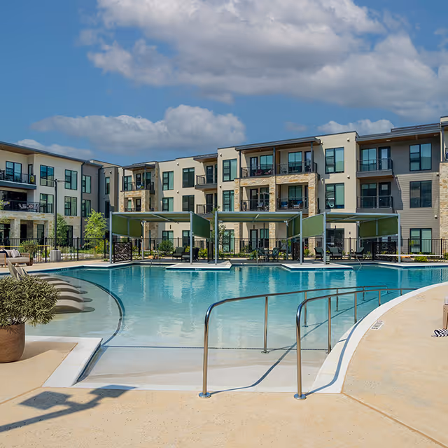 Outdoor swimming pool area with lounge chairs partially submerged in the water, shaded cabanas, and a multi-story residential building in the background under a partly cloudy sky.