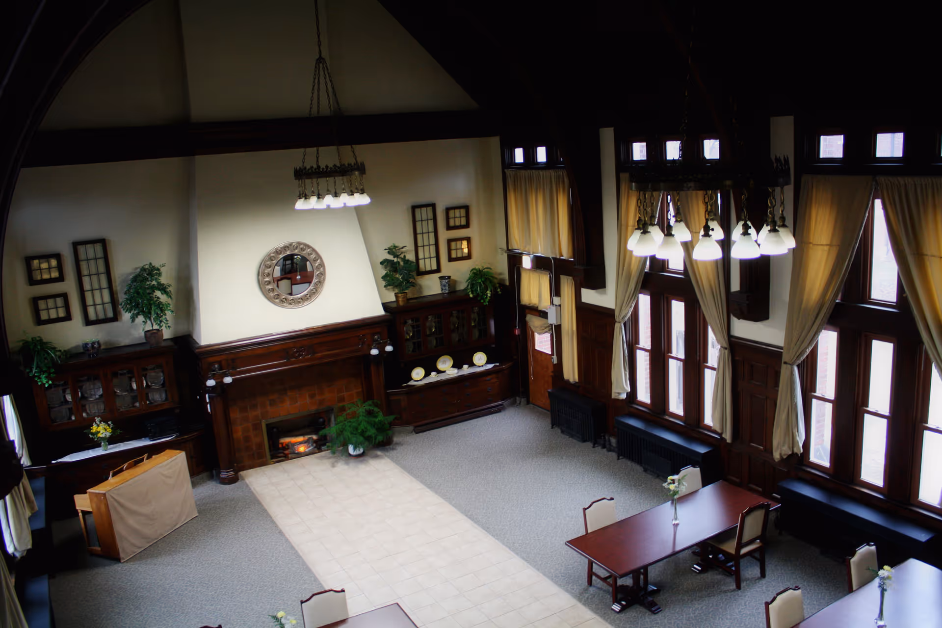 A spacious, high-ceilinged room with large windows covered by beige curtains, wooden furniture including tables and chairs, a fireplace with a decorative round mirror above it, and plants placed on cabinets and near the fireplace. The room has a mix of carpet and tile flooring and is illuminated by hanging light fixtures.