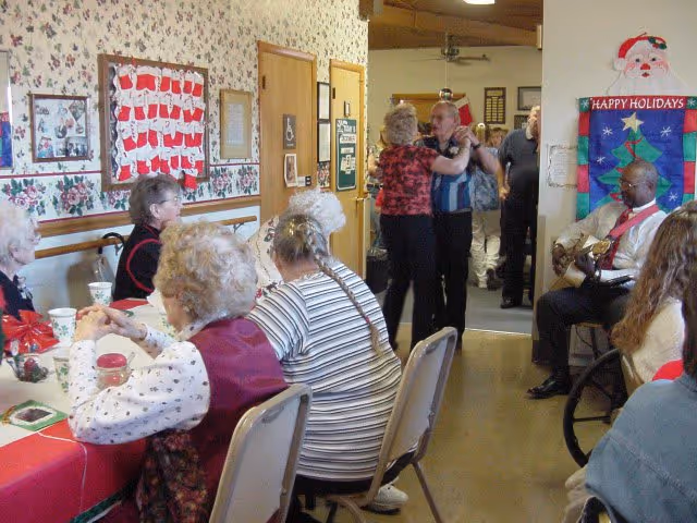 A group of elderly people gathered in a decorated room with holiday-themed decorations. Some are seated at a table covered with a red tablecloth, while others are dancing in the background. A man is playing a guitar near a wall decorated with a 'Happy Holidays' banner featuring a Christmas tree and Santa Claus.