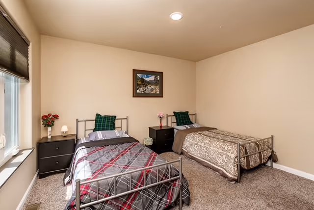 A bedroom with two twin metal-frame beds, nightstands, lamps, a window, and a framed picture on the wall.