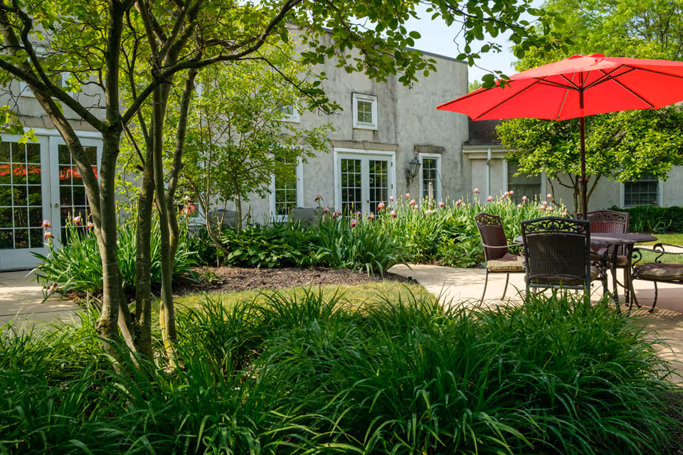 Outdoor patio area at New Albany - An Optalis Health & Rehabilitation Center featuring a round table with four chairs and a large red umbrella. The patio is surrounded by lush green plants, trees, and flowering bushes, with the building's exterior and glass doors visible in the background.